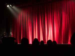 A spotlight shines on the middle of a stage. The red theatre curtains are drawn ready for a show to begin.