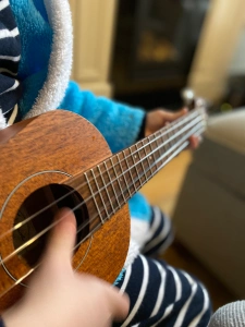 A small child, dressed in stripy pyjamas and a fluffy blue dressing gown is playing a brown, wooden ukulele.